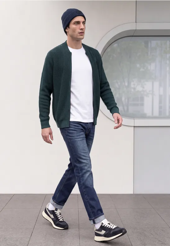 Ein Mann in Strickjacke, T-Shirt, Jeans und Mütze geht vor moderner Fassade.