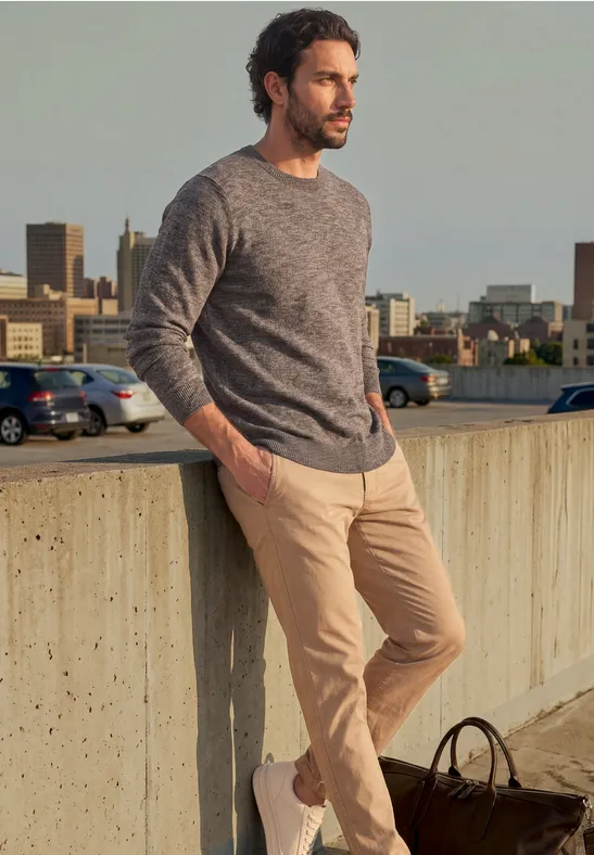 Ein Mann lehnt entspannt an einer Betonmauer, trägt Pullover und Chinohose, mit Tasche am Boden, Stadt im Hintergrund.