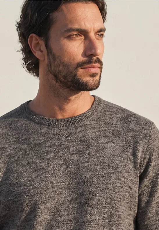 Ein Mann mit dunklen Haaren und kurzem Bart trägt einen grauen Strickpullover und blickt nach rechts.