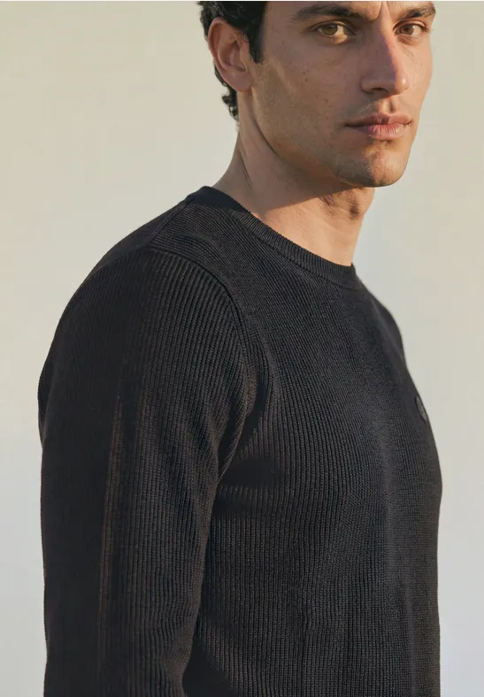 Ein Mann in schwarzem Pullover blickt seitlich in die Kamera vor neutralem hellem Hintergrund.