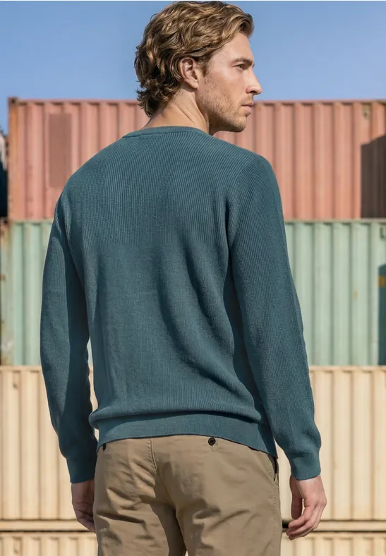 Ein Mann in grünem Pullover und beigefarbenen Hosen steht vor bunten Frachtcontainern.