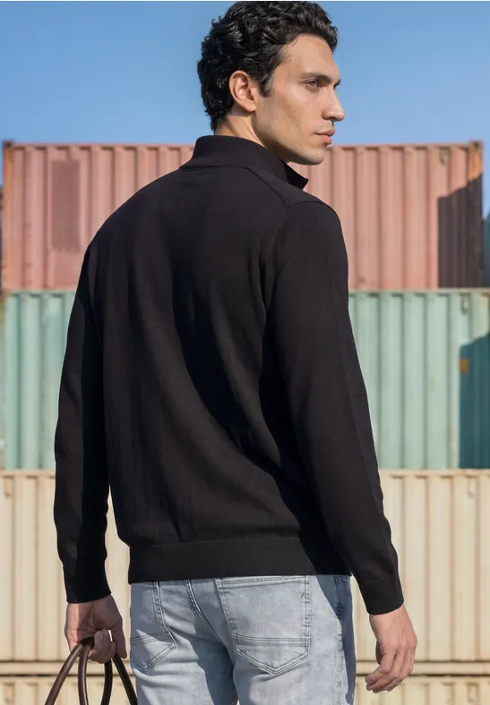 Ein Mann steht in schwarzem Pullover und hellen Jeans vor bunten Containern und hält eine Tasche.