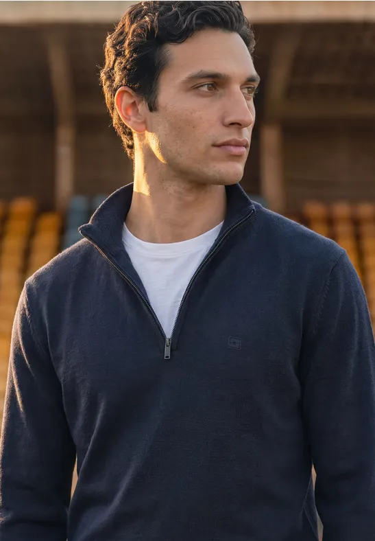 Junger Mann in dunkelblauem Pullover mit halbem Reißverschluss und weißem T-Shirt vor unscharfem Hintergrund.