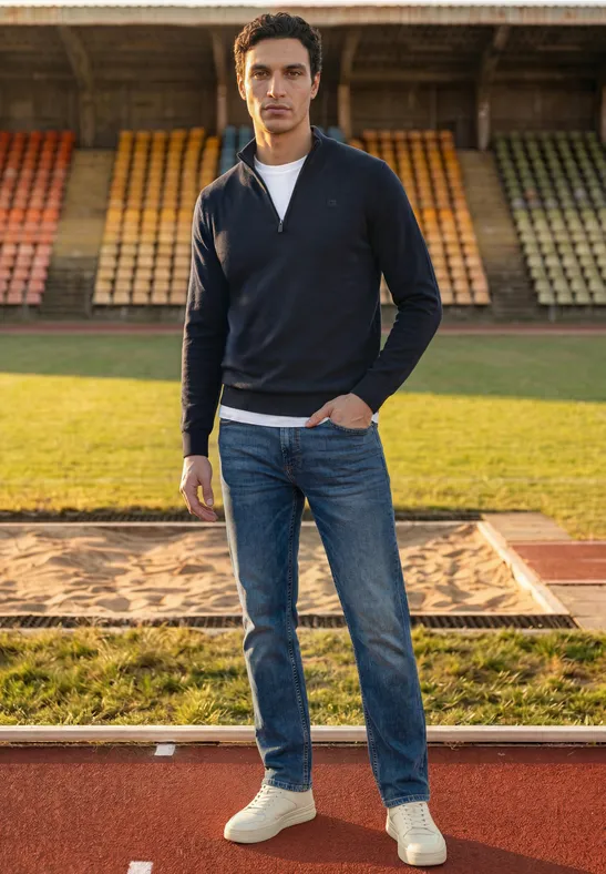 Ein junger Mann in Jeans, Sneakern und Pullover steht selbstbewusst auf einer Laufbahn vor Tribünen.