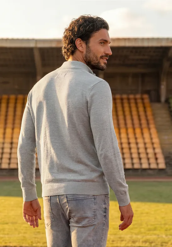 Een man in een grijze trui en spijkerbroek staat op een sportveld en kijkt over zijn schouder.
