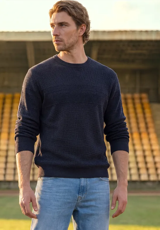 Ein Mann in dunklem Pullover und Jeans steht vor leeren Tribünen im Freien.