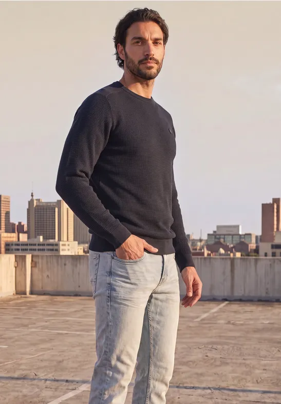 Ein Mann im schwarzen Pullover und Jeans steht auf einem Dach mit Stadtsilhouette im Hintergrund.