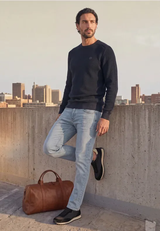 Mann in lässigem Pullover und Jeans lehnt auf Dachterrasse mit Stadtkulisse im Hintergrund.