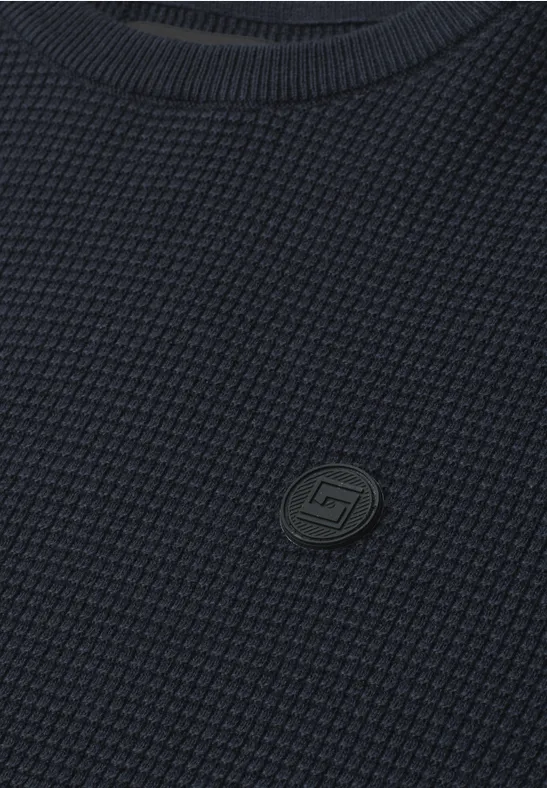 Nahaufnahme eines dunkelblauen, waffelgestrickten Pullovers mit rundem Markenlogo.