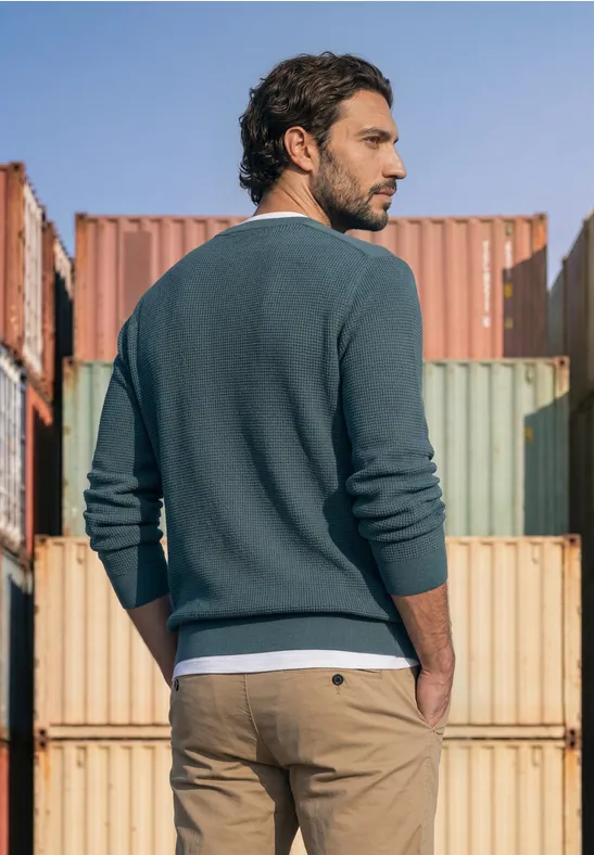 Ein Mann in Pullover und Chino steht vor bunten Frachtcontainern und blickt seitlich.