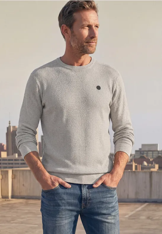 Ein blonder Mann mit grauem Pullover und Jeans steht lässig auf einem Parkdeckdach und schaut in die Ferne.