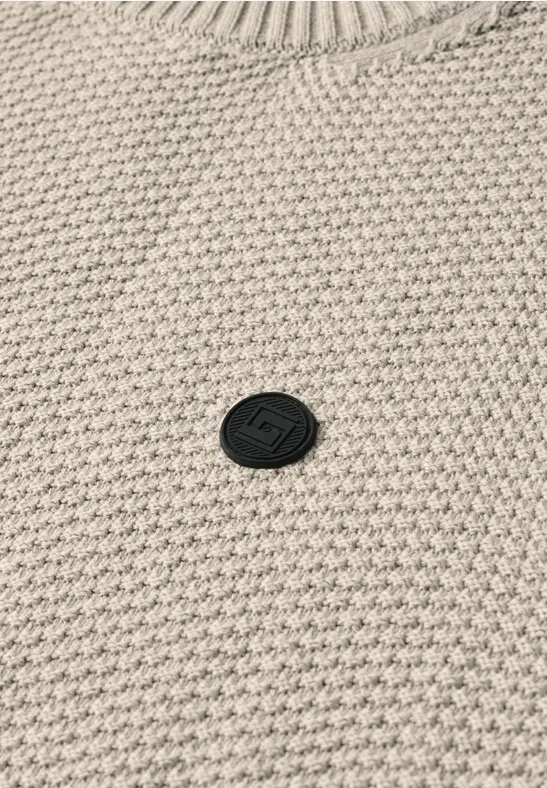 Beżowy sweter z waflowym splotem, z małym, okrągłym czarnym logo na środku.