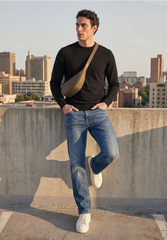 Een man in zwarte trui, jeans en witte sneakers leunt relaxt tegen een betonnen rand met stadszicht.