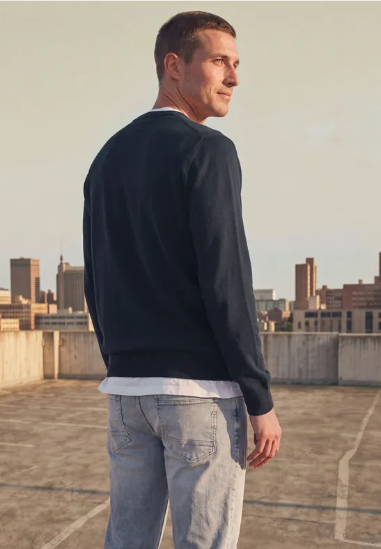 Man staat op een parkeerdek, draagt een donkerblauwe trui, wit shirt en spijkerbroek, kijkt over zijn schouder.