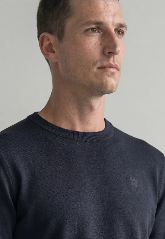 Portret van een man in een donkerblauwe trui, in profiel tegen een neutrale achtergrond.