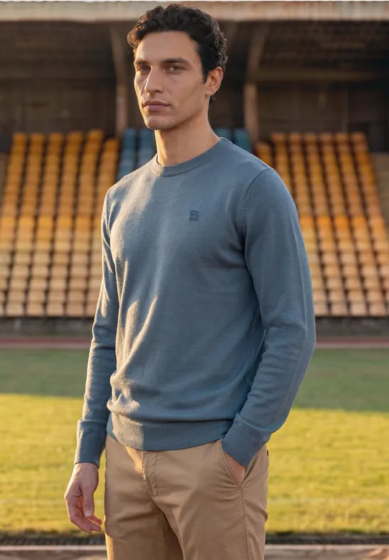 Mann im hellblauen Pullover und beigen Hosen vor leeren Tribünen.