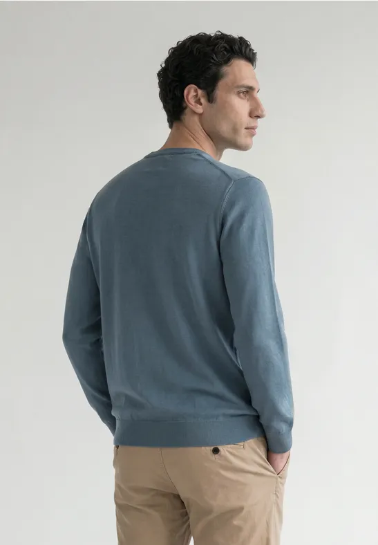 Ein Mann im blau-grauen Pullover und beiger Hose steht seitlich, eine Hand in der Tasche.