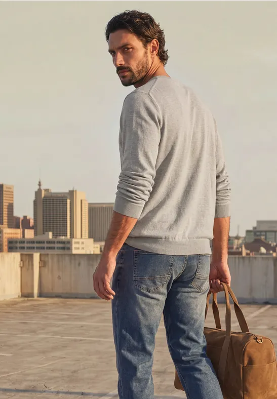 Mann steht auf einem Dach, blickt zurück, trägt einen grauen Pullover, Jeans und hält eine Reisetasche.