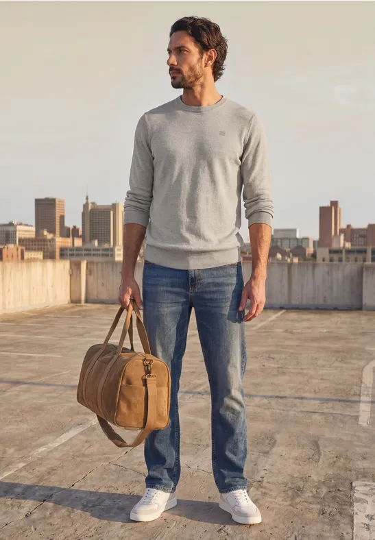 Mann im Pullover, Jeans und Sneakers, trägt Reisetasche auf Dach mit Stadtblick.