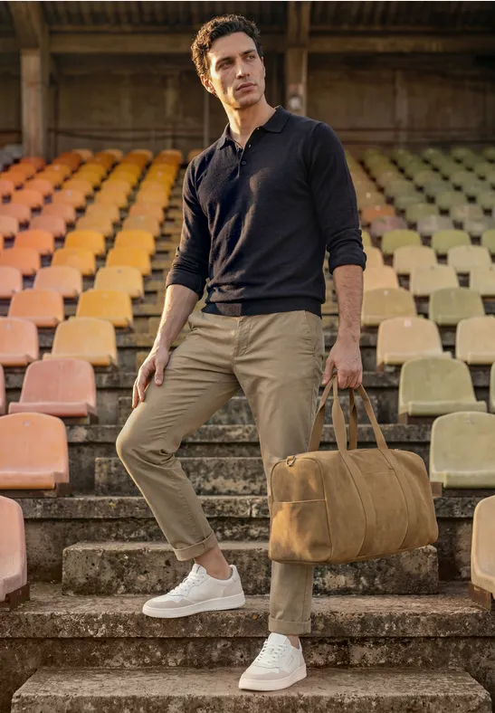 Een man staat op stadiontrappen met gekleurde stoelen, draagt vrijetijdskleding en houdt een beige reistas bij zich.