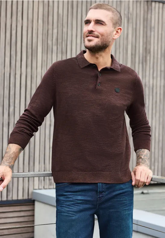 Mann trägt braunen Pullover mit Knopfleiste und blaue Jeans, steht lässig vor Holzlamellen