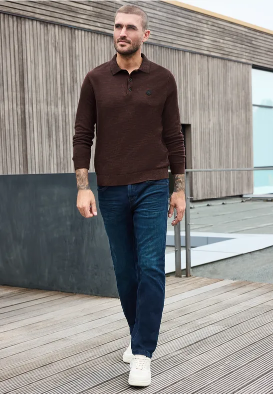 Ein Mann in braunem Pullover, Jeans und weißen Sneakern läuft auf einer Holzterrasse vor modernem Gebäude.