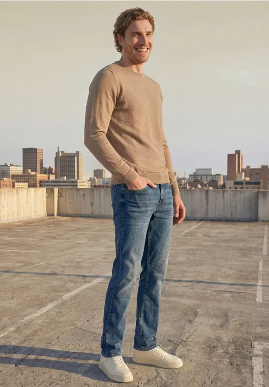 Ein lächelnder Mann in Jeans und beigem Pullover steht auf einem Parkdeckdach mit Stadtblick.