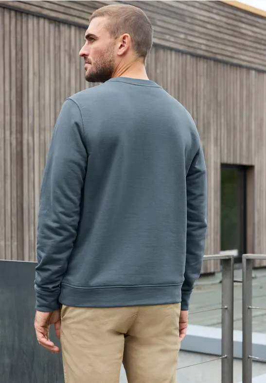 Ein Mann in graublauem Sweatshirt und beiger Hose steht seitlich vor einem Holzgebäude.