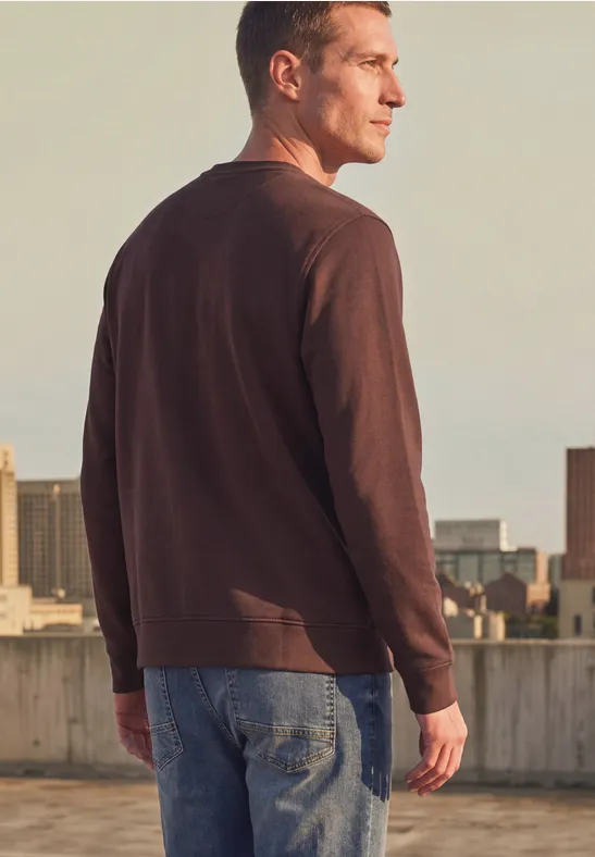 Mann in braunem Langarmshirt und Jeans steht auf einem Dach und schaut auf die Stadt.