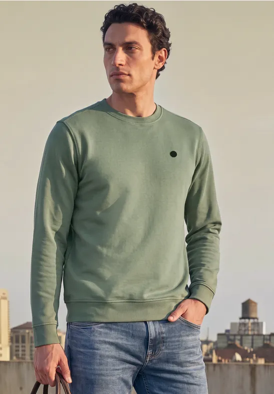 Un homme porte un pull vert et un jean, debout devant une ville au coucher du soleil.