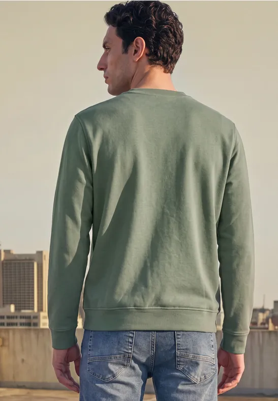 Un homme sur un toit, dos à l'appareil, porte un sweat vert et un jean devant une skyline urbaine.