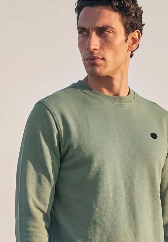 Un homme aux cheveux bouclés porte un sweat vert clair à col rond et design épuré.