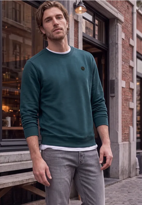 Ein Mann in grünem Pullover und grauen Jeans steht entspannt vor einem Laden auf Kopfsteinpflaster.
