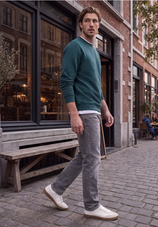 Ein Mann mit grünem Pullover, grauer Hose und weißen Sneakers blickt in der Altstadt vor einem Caféfenster.