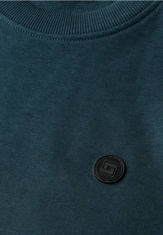 Dunkelgrünes Shirt mit rundem, schwarzem Logo-Aufnäher an der Brust.