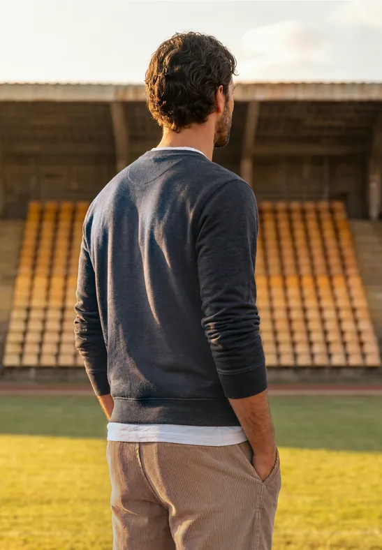 Ein Mann im Pullover steht auf einem Stadionrasen und blickt auf leere Tribünen.