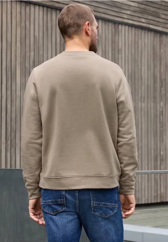 Ein Mann von hinten in graubraunem Pullover vor holzvertäfelter Fassade.