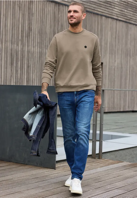 Ein Mann läuft in Jeans, beigem Pullover und weißen Sneakern und trägt eine Jacke über dem Arm.
