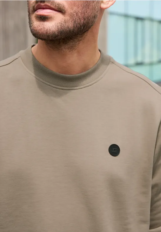 Nahaufnahme eines Mannes trägt ein taupefarbenes Shirt mit Rundhalsausschnitt und kleinem schwarzem Logo.