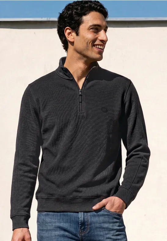 Ein lächelnder Mann in dunklem Pullover mit Reißverschluss und Jeans vor weißem Hintergrund.