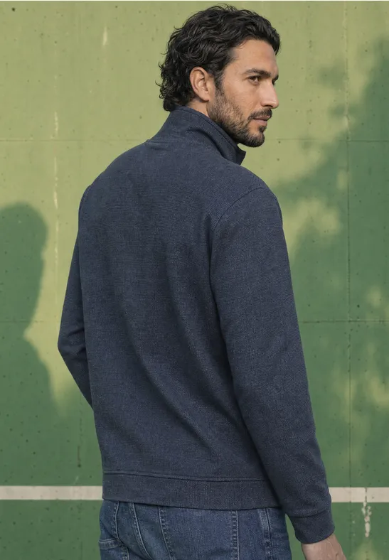 Ein Mann in dunkelblauem Pullover steht vor grüner Wand und schaut seitlich über die Schulter.