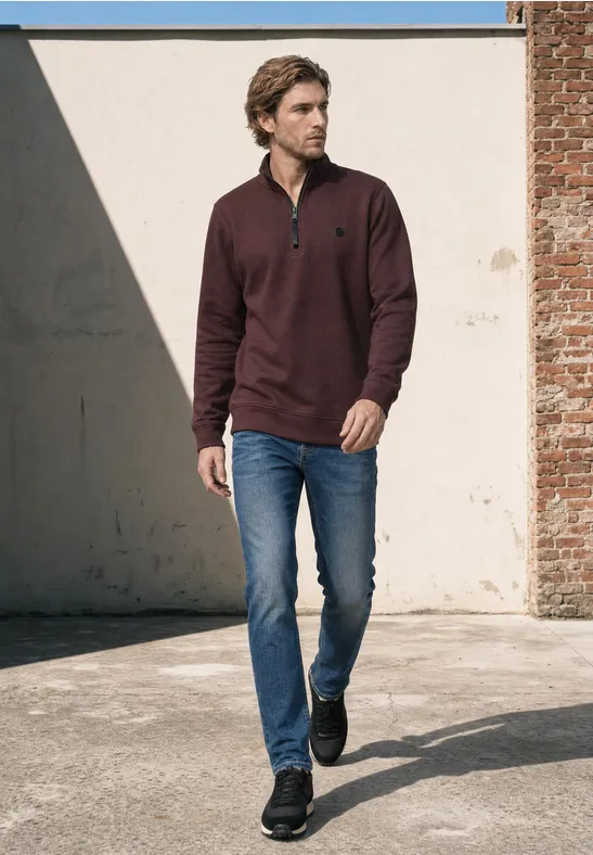 Ein Mann spaziert an einer Wand entlang, trägt dunkelroten Pullover, blaue Jeans und schwarze Sneakers.