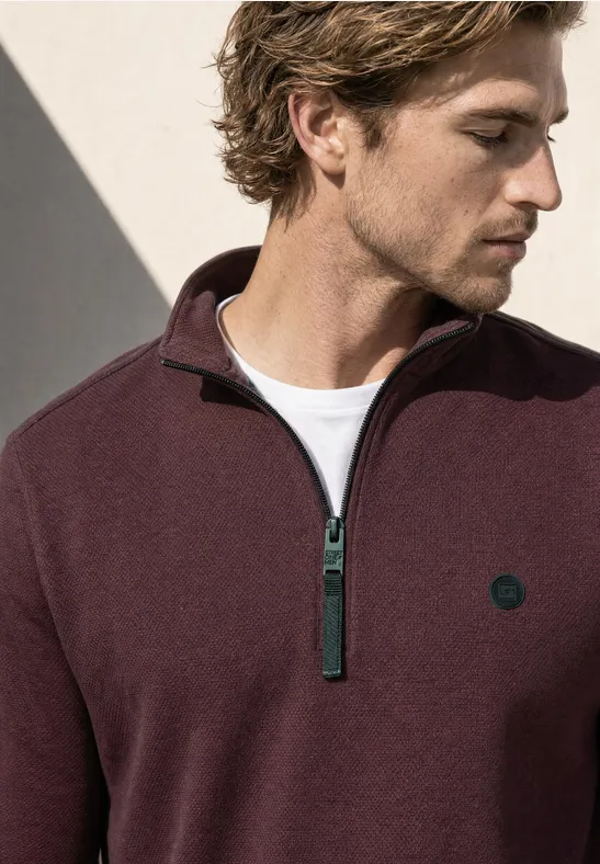 Ein Mann in einem burgunderroten Pullover mit Reißverschluss und kleinem Logo, der nach unten blickt.