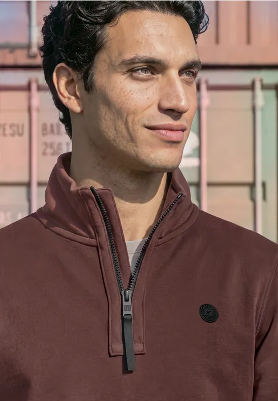 Ein Mann in einem dunkelroten Zip-Pullover vor bunten Containern.