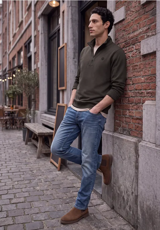 Ein Mann lehnt lässig an einer Backsteinwand in einer Kopfsteinpflastergasse, trägt Pullover und Jeans.