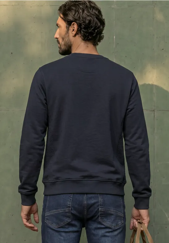 Ein bärtiger Mann im dunkelblauen Pullover und Jeans steht mit dem Rücken zur Kamera, hält eine braune Tasche.