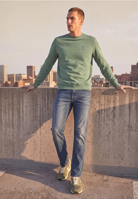 Een man in een groen sweatshirt en spijkerbroek staat op een dak en kijkt over de stad.