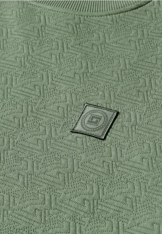 Groene stof met doorlopend geometrisch patroon en een klein vierkant zwart logo-patch.