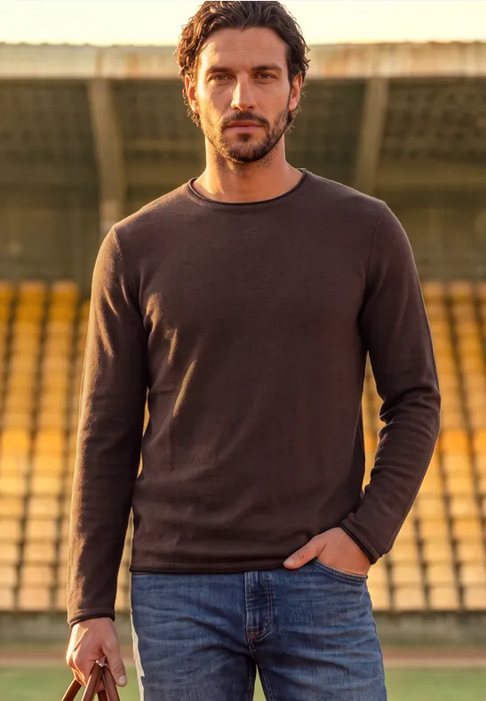 Ein Mann in braunem Pullover und Jeans steht vor leeren Stadionrängen.