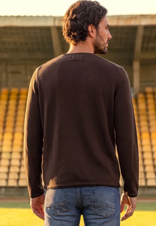 Mann mit braunem Pullover und Jeans steht mit dem Rücken zur Kamera in einem Stadion mit Sitzreihen.